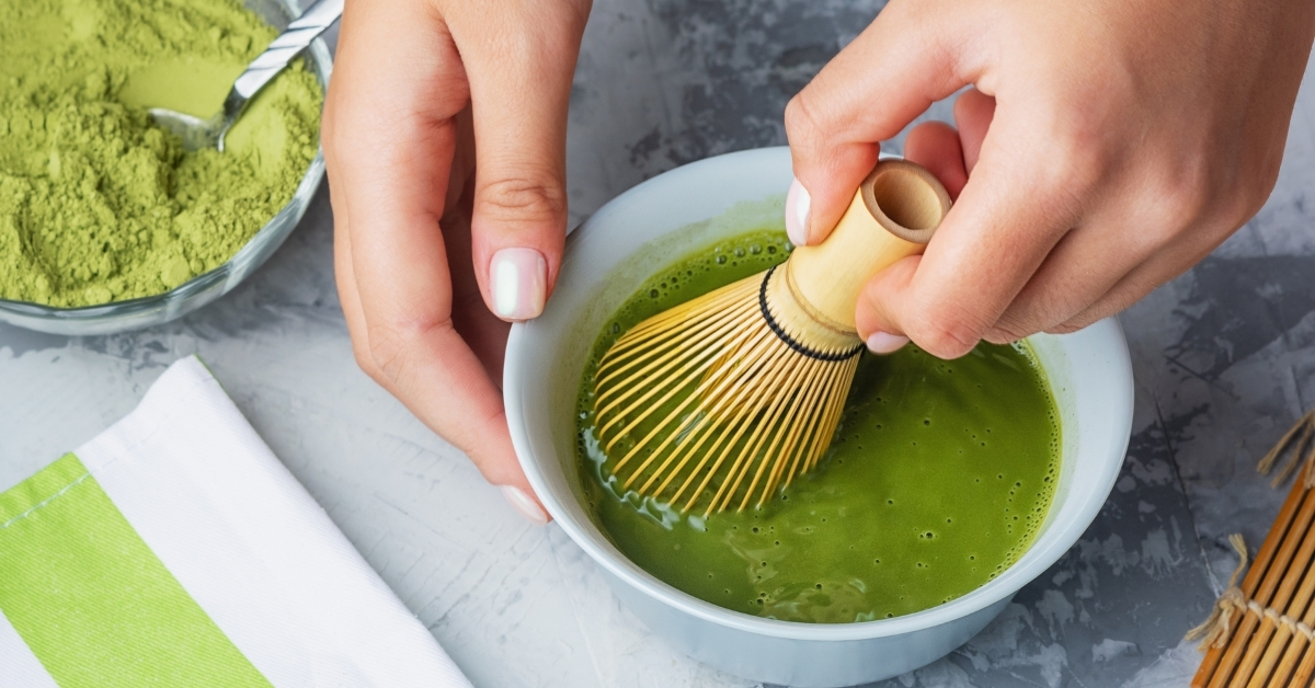 Kéz bambusz habverővel matchát készít egy tálban, a matcha készítésének lépéseit bemutató cikkhez